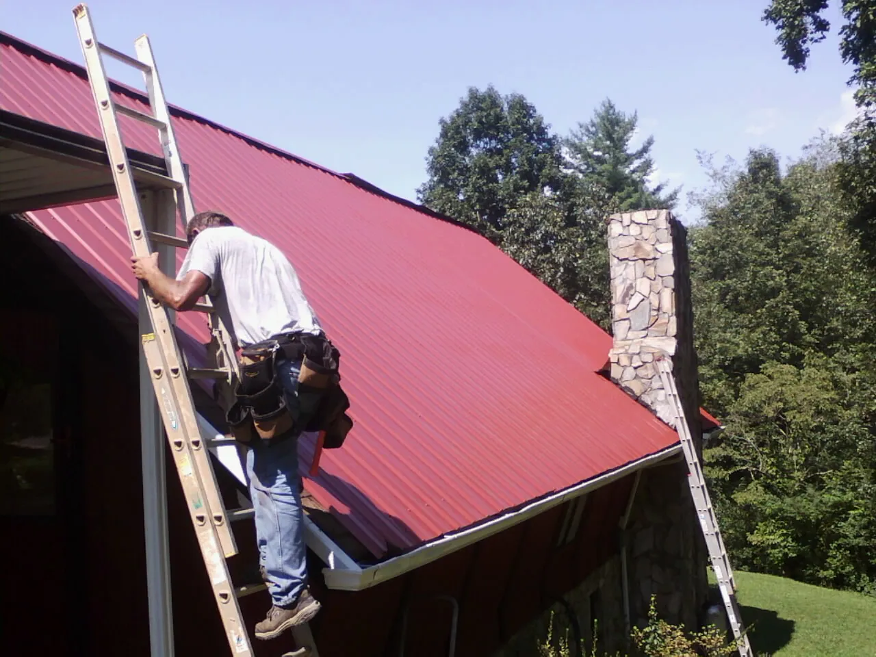 Roofer on a ladder beside a red metal roof and stone chimney — Soffit & Fascia Repair by Roofing Brothers in Mount Aetna